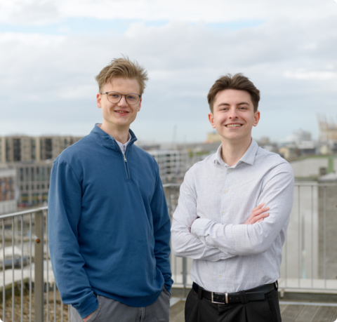 Zwei junge Männer lächeln draußen an eine Brüstung gelehnt in die Kamera, im Hintergrund ist Wasser und ein verschwommen ein Schiff sowie ein segelartiges Gebäude zu sehen | © Antje Schimanke